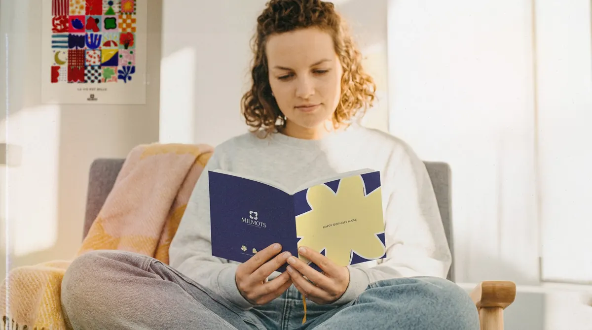 Une femme lit un carnet Milmots à couverture bleue et jaune, devant une affiche de carrés illustrés.